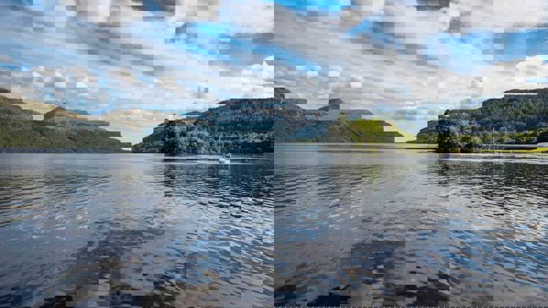 A large body of water surrounded by mountains and hills under a cloudy blue sky.