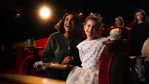 A smiling adult and a young child enjoy a movie in a dimly lit cinema, sharing food and drinks from their plush red seats.