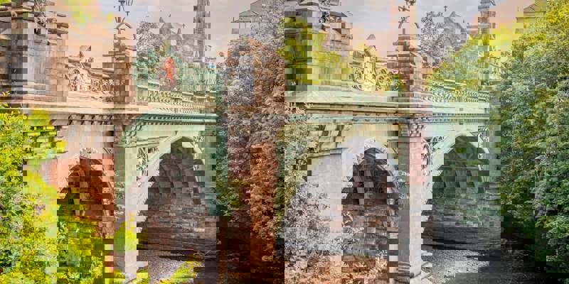 A picturesque stone and iron bridge over a river, with two people walking on the riverside path and a church spire rising behind trees in the distance.