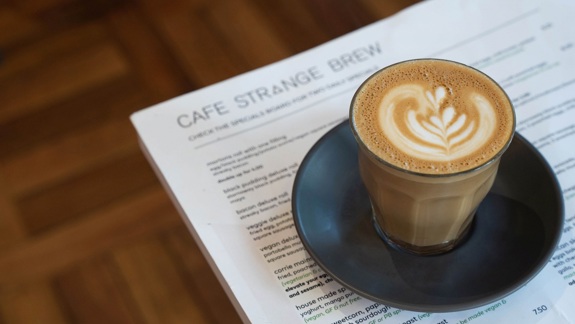A close-up of a velvety latte on a black saucer, sitting on a menu from "Cafe Strange Brew."