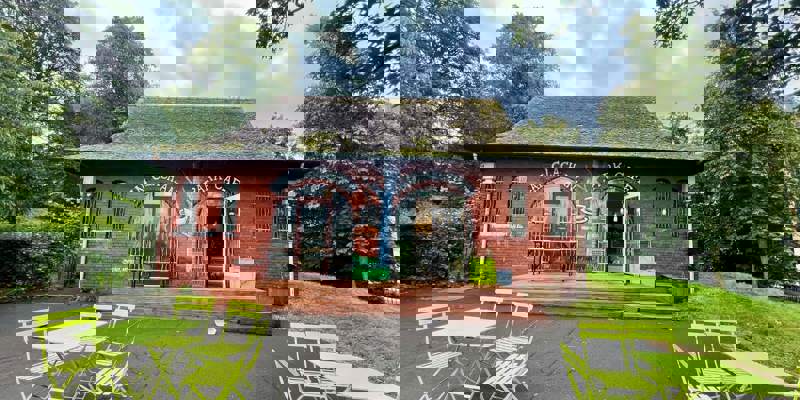 A small red-brick café building with outdoor seating featuring bright green tables and chairs, surrounded by trees.