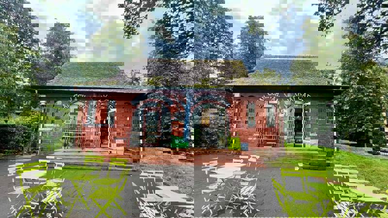 A small red-brick café building with outdoor seating featuring bright green tables and chairs, surrounded by trees.