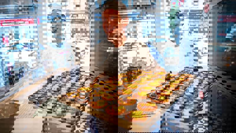A baker in a white uniform smiles while proudly holding a large tray of freshly baked, traditional Portuguese Pastéis de Nata in a bakery.