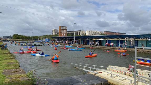 A wide shot of Glasgow's Pinkston Watersports centre, showing dozens of people in colourful kayaks and canoes paddling on a canal under a cloudy sky.