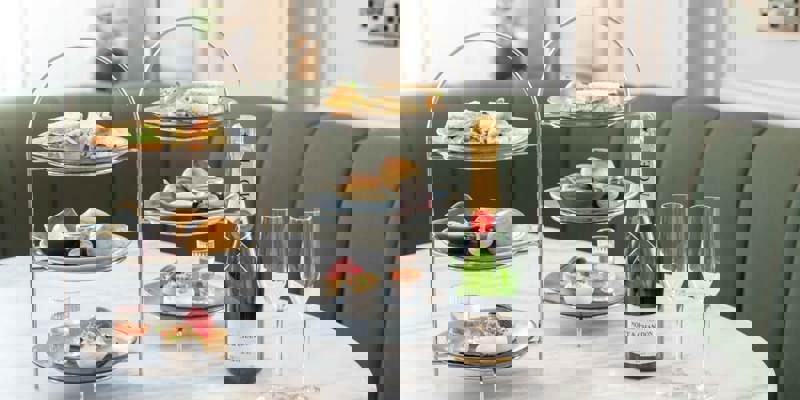 A luxury afternoon tea set on a marble table with two three-tiered stands of sandwiches, scones, and pastries, next to a bottle of Moët & Chandon.