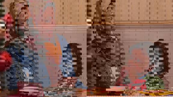 A woman smiling at a child sitting opposite her at a table with a pizza and toys, in a warm, relaxed restaurant setting.