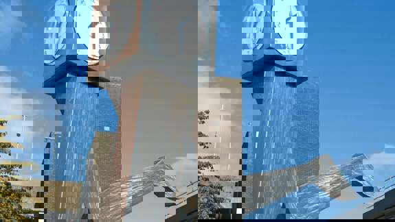 A unique modern sculpture features a large, metallic abstract figure with clocks on its head, seemingly in motion, set against a clear blue sky.