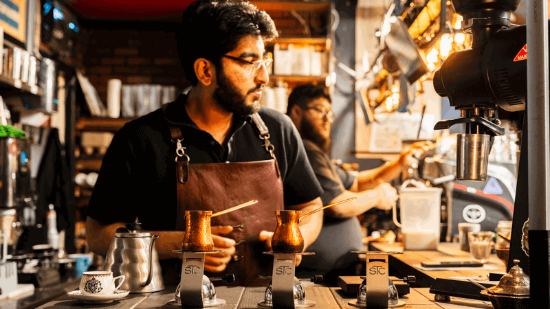 Barista in brown leather apron prepares drinks with copper pots on burners, surrounded by coffee equipment in cosy shop with shelves and warm lighting.