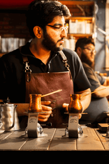 Barista in brown leather apron prepares drinks with copper pots on burners, surrounded by coffee equipment in cosy shop with shelves and warm lighting.