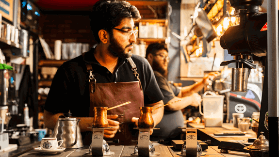 Barista in brown leather apron prepares drinks with copper pots on burners, surrounded by coffee equipment in cosy shop with shelves and warm lighting.