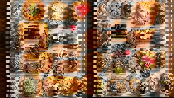 A flat lay of assorted sweet and savoury bakery treats on a tray, including croissants, cookies, brownies, and tarts.