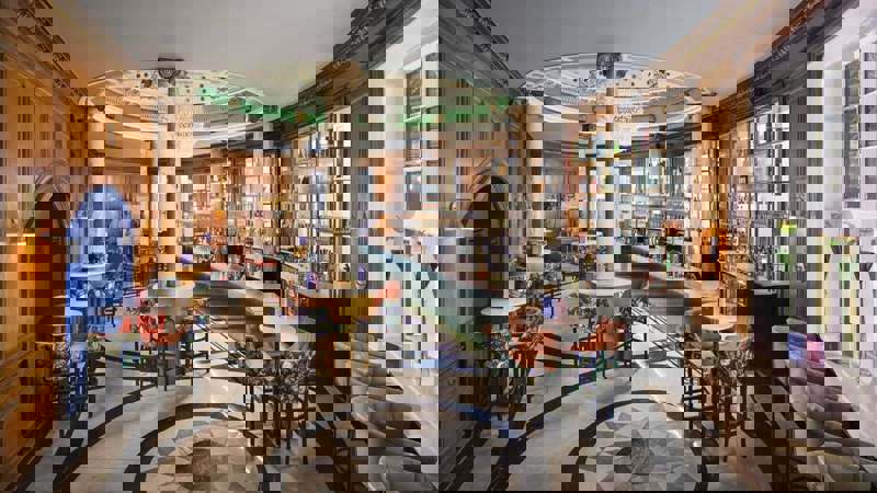 A grand hotel bar with high ceilings, ornate chandeliers, large arched windows, and a mix of velvet and leather seating, overlooking a busy train station concourse.