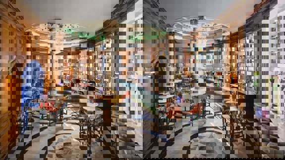 A grand hotel bar with high ceilings, ornate chandeliers, large arched windows, and a mix of velvet and leather seating, overlooking a busy train station concourse.