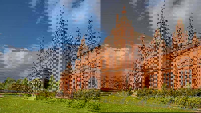 A large red sandstone building with towers, arches, and green lawns in front.