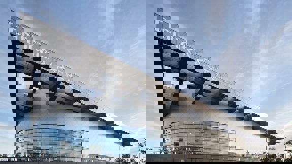 The modern, angled facade of the Emirates Arena, a large sports venue with extensive glass windows under a blue sky with clouds.