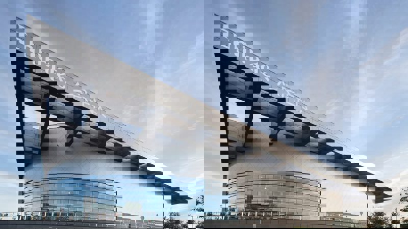 The modern, angled facade of the Emirates Arena, a large sports venue with extensive glass windows under a blue sky with clouds.