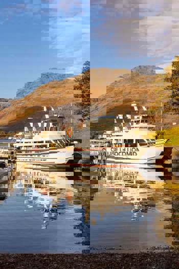 A scenic view of a "Cruise Loch Lomond" boat docked on a serene loch, with majestic mountains and a lone tree with autumn foliage in the background under a blue sky.