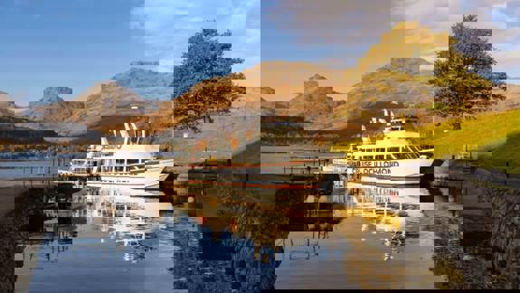 A scenic view of a "Cruise Loch Lomond" boat docked on a serene loch, with majestic mountains and a lone tree with autumn foliage in the background under a blue sky.
