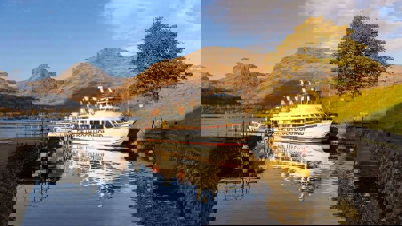 A scenic view of a "Cruise Loch Lomond" boat docked on a serene loch, with majestic mountains and a lone tree with autumn foliage in the background under a blue sky.