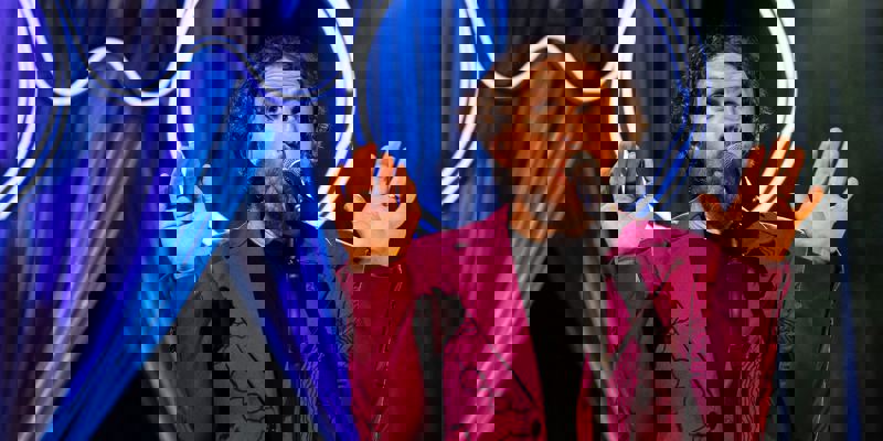 Man with curly hair in a magenta jacket with black line drawings performing on a stage.