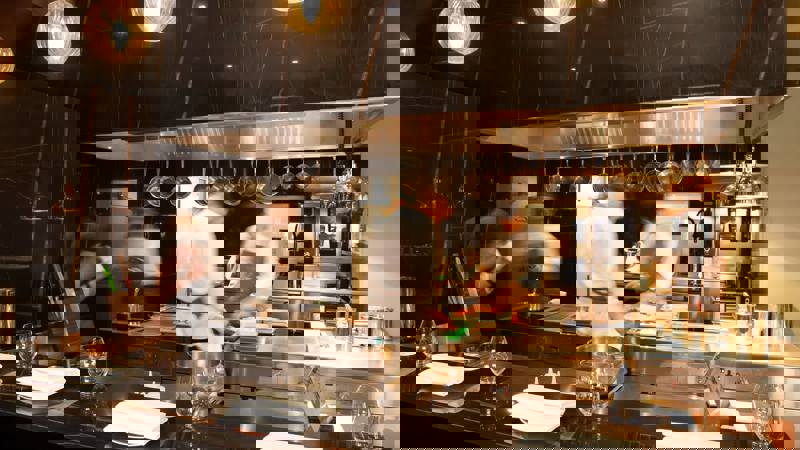 Chefs work in a bustling, modern kitchen with a black marble countertop and hanging lights.