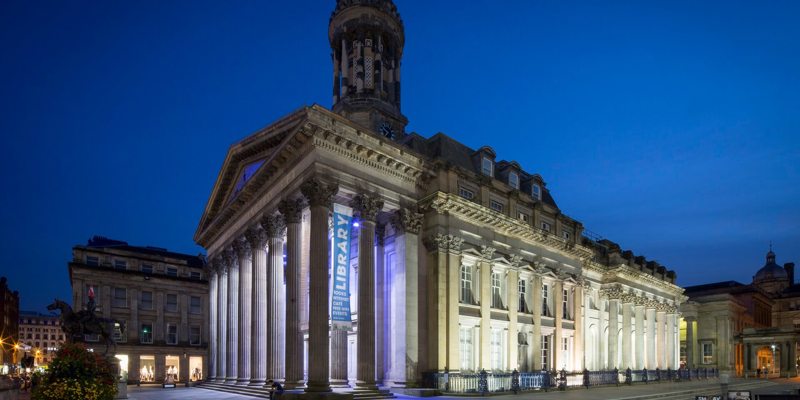 The Gallery of Modern Art (GOMA) at night, with its classical facade and columns dramatically lit against a dark blue sky.