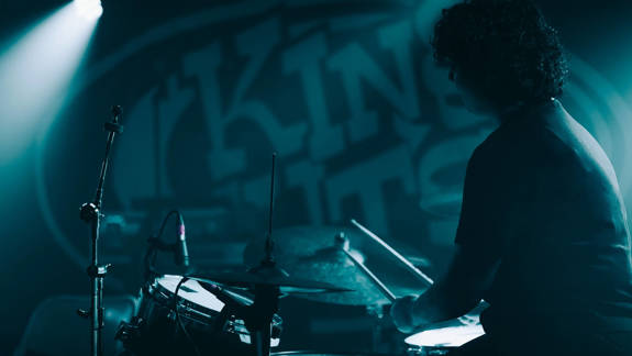 A drummer is actively playing a drum kit on a dimly lit stage, with a "King Tut's Wah Wah Hut" logo visible in the blurred background.