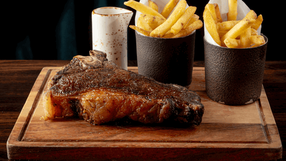 Juicy, cooked steak with seared crust on wooden board, served with golden French fries in cups and a white sauce container in the background.