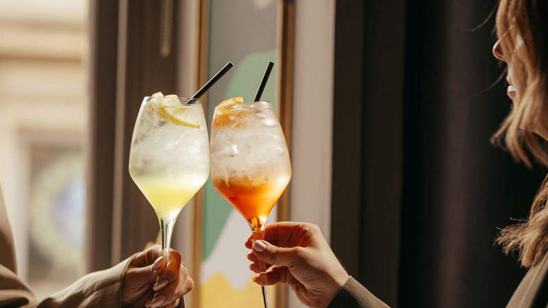 Two people in beige blazers clink glasses holding light yellow and orange cocktails in a sunlit interior.