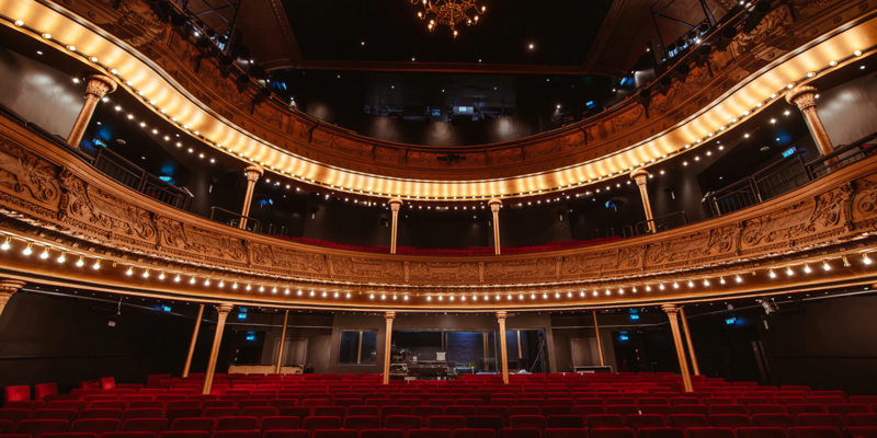 Interior of Citizens Theatre in Glasgow featuring ornate balconies, soft, glowing lights, red velvet upholstered seating, and a grand chandelier above the audience seating. 