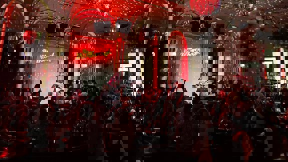 A live music venue is bathed in red light from disco balls and stage lights, where a musician plays guitar to a seated audience.