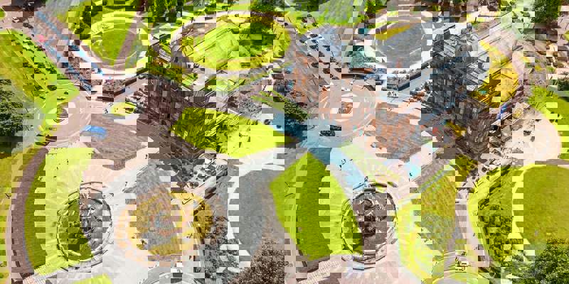 An aerial view of a large, brown brick building with a glass roof surrounded by a series of circular green lawns and paved paths.