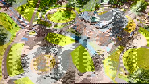 An aerial view of a large, brown brick building with a glass roof surrounded by a series of circular green lawns and paved paths.