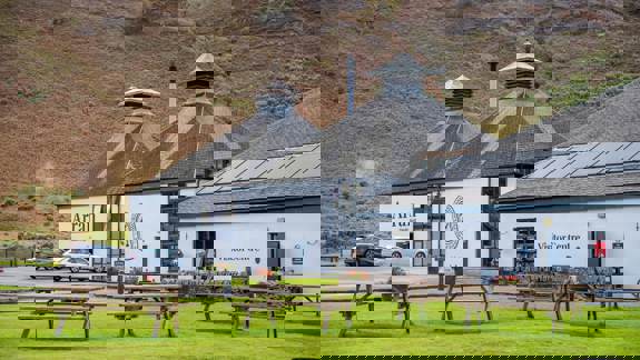 The white-walled Lochranza Distillery featuring traditional pagoda roofs, set against a steep hillside with wooden picnic benches on a green lawn in the foreground.