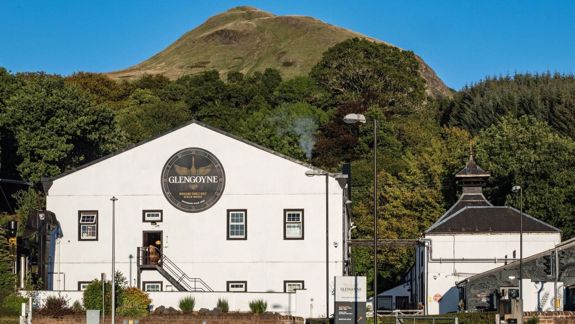 A sunny view of the Glengoyne Distillery, a white building nestled against a green, tree-covered hill with a large hill behind it.