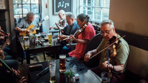 Group of musicians playing acoustic guitars and fiddles around a table with drinks inside a cozy pub, with a chalkboard sign reading ‘Failte Towers’ behind them in the background.