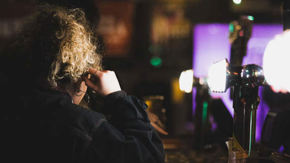 Two people talk seated at a dimly lit bar with illuminated beer taps and hanging lights, creating a chilled, intimate atmosphere.