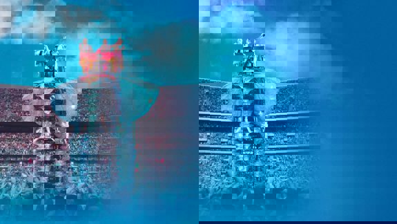 Take That performs on an enormous, glittering elephant stage prop for a huge, cheering crowd in a stadium setting under a blue sky.