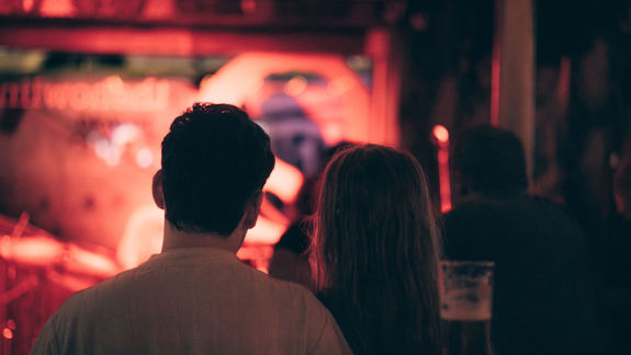Couple enjoying live music at a warm, vibrant bar with a red-lit stage and musicians visible in background.