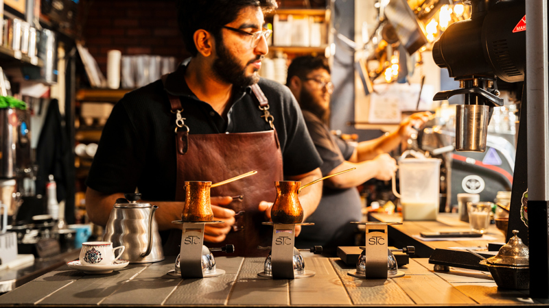 A barista in a leather apron prepares traditional coffee using copper cezves on heated sand. The background shows a busy, warm-lit cafe interior with professional brewing equipment.