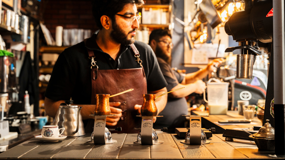 A barista in a leather apron prepares traditional coffee using copper cezves on heated sand. The background shows a busy, warm-lit cafe interior with professional brewing equipment.