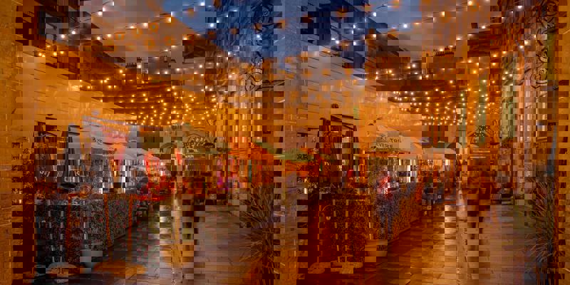 An outdoor dining area at night, with strings of lights illuminating a cobbled courtyard with a few people walking through.