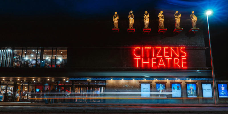 Exterior of Citizens Theatre in Glasgow at night with bright red neon sign, illuminated statues on the roof, and crowds visible through large windows on the left.