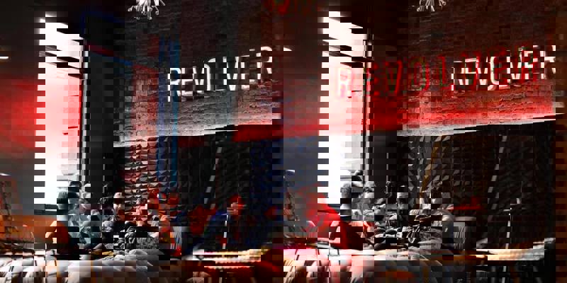 Dark, trendy hotel lobby with a neon "REVOLVER" sign, exposed brick, and a group of people relaxing on large, cushioned seating.