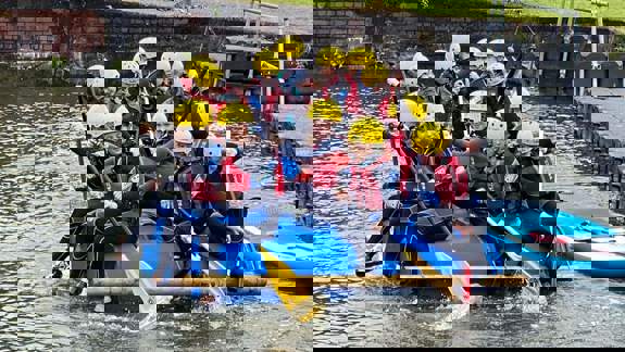 A group of children wearing yellow helmets and wetsuits, paddling a small, makeshift raft on a body of water.