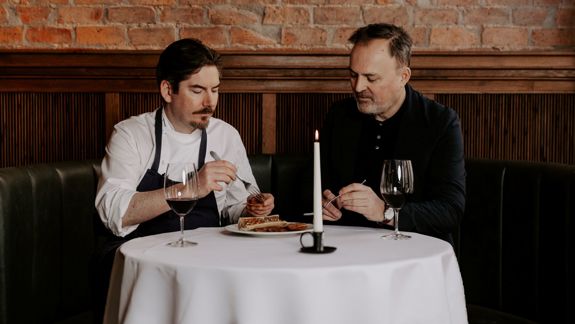 Two men seated at a restaurant table tasting a dish, with wine glasses and a candle, against a brick wall backdrop.