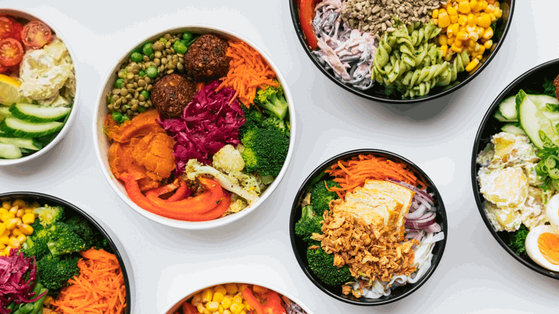 Colorful healthy food bowls featuring fresh vegetables, grains, beans, falafel, pasta, carrots, broccoli, and cherry tomatoes on a white background.