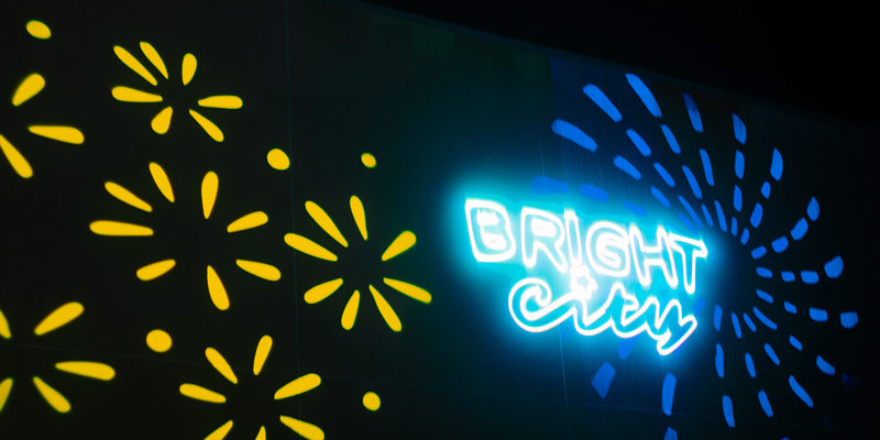 Neon sign saying "BRIGHT CITY" glowing blue and white on a dark wall, flanked by projected yellow and blue firework-like patterns.