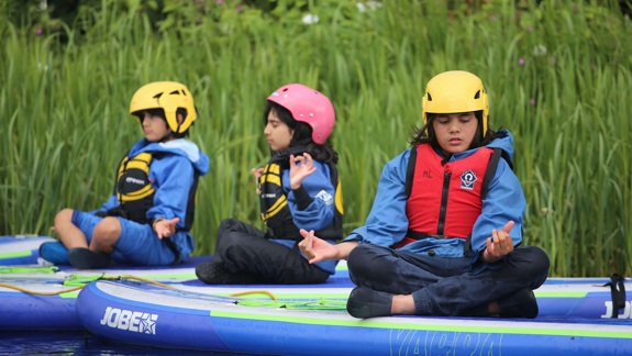 Three young people sit on paddleboards on a body of water, wearing life vests and helmets.