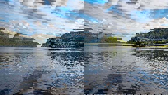 A view of a beautiful loch with mountains on either side under a blue sky with white clouds.